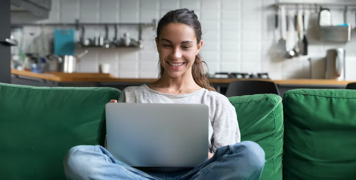 A blonde-haired woman sitting cross-legged on a couch, smiling at her open laptop.