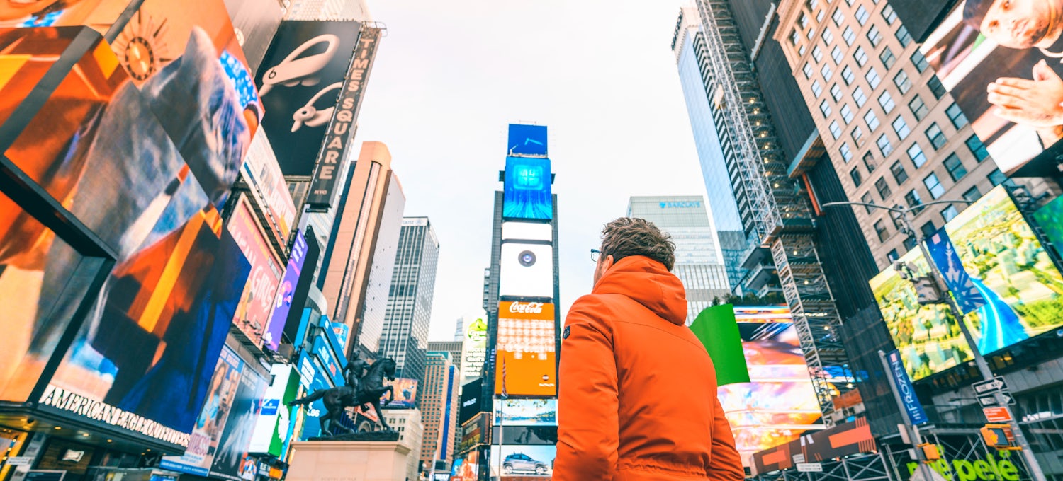 [Imagen destacada] Un hombre con una parka naranja mira los anuncios en Times Square.