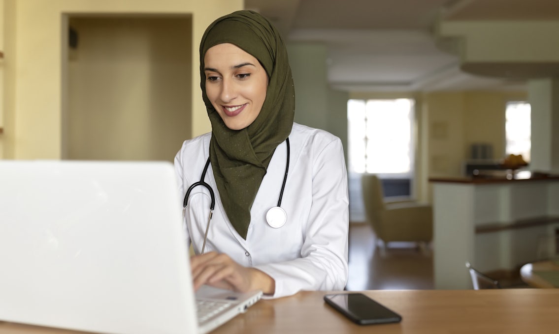 A woman wearing a hijab, a white lab coat, and a stethoscope sits using a laptop.  