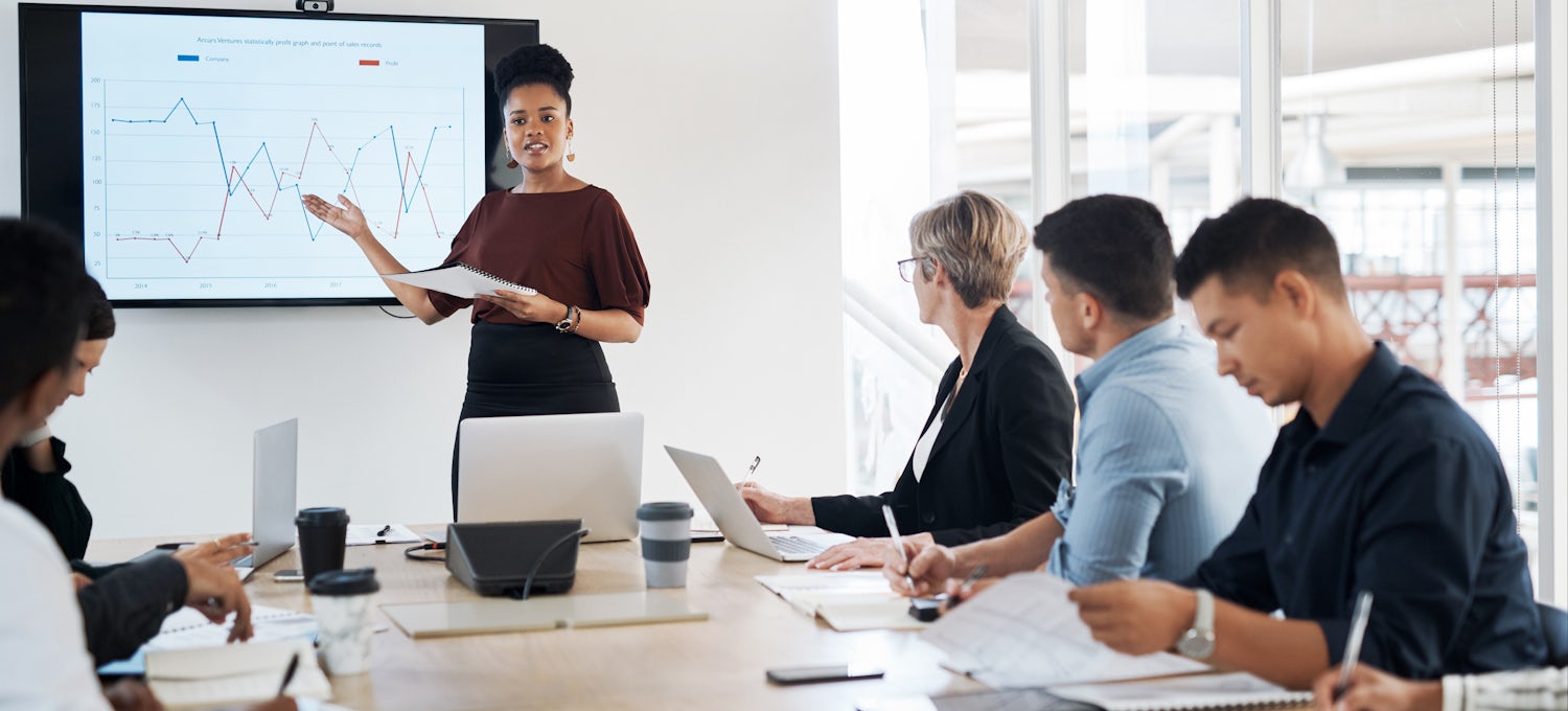 [Imagen destacada] Una mujer se coloca delante de una pizarra digital y dirige una reunión de estrategia de marketing con varios compañeros de trabajo. 