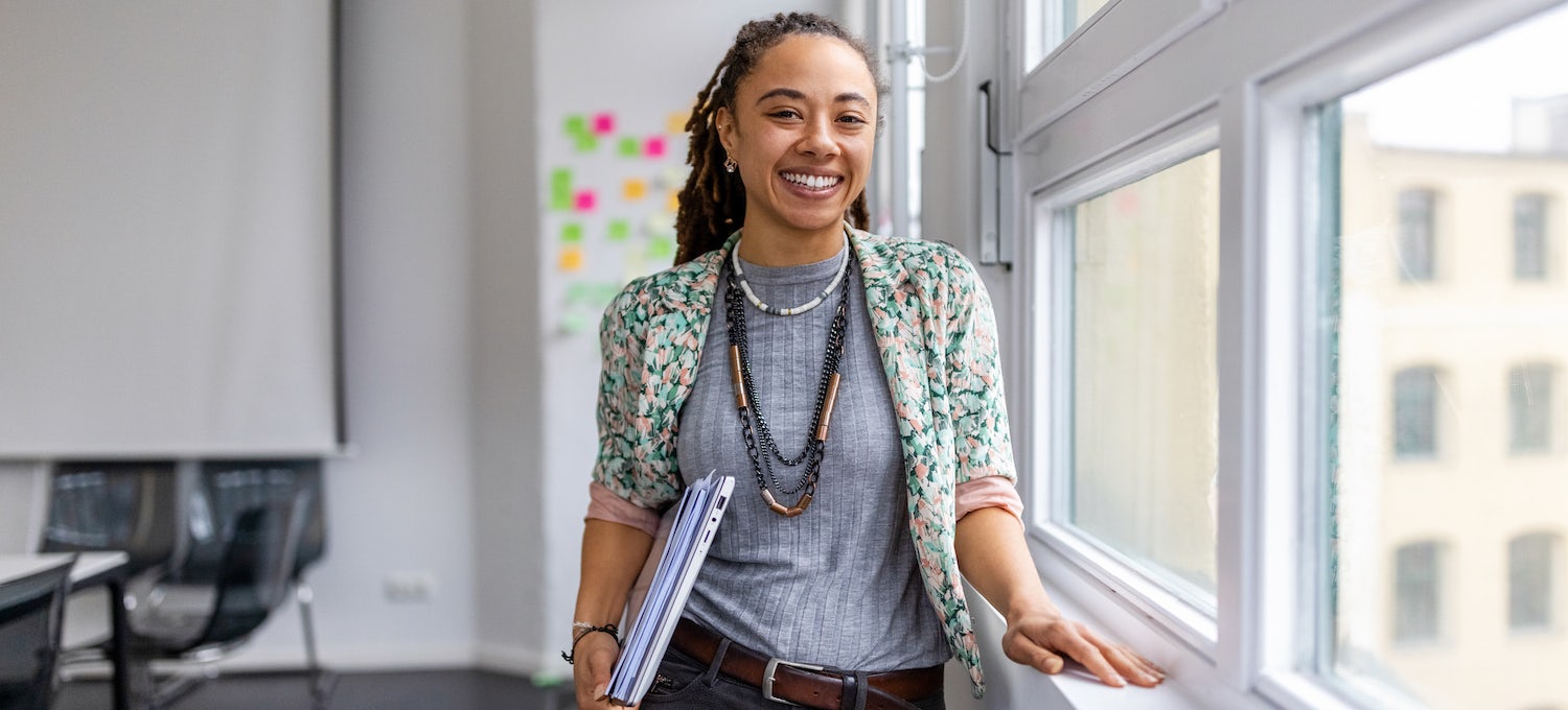 [Featured image] A woman of color carries a laptop and some papers. She stands in front of an office window smiling. 