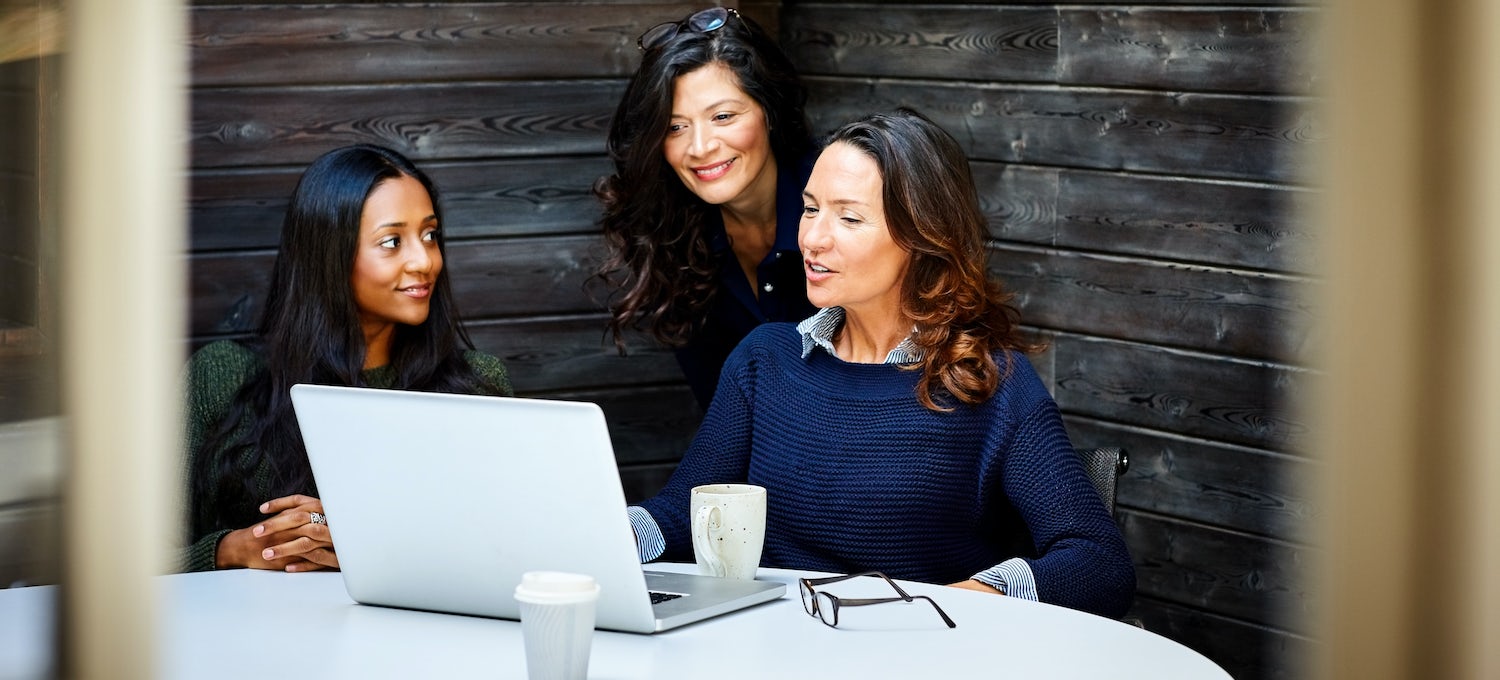 A marketing manager meets with her team at a conference table. One woman has a laptop, glasses, and a coffee cup in front of her.
