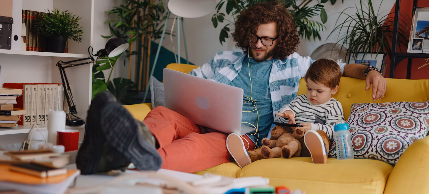 A man sits on a yellow sofa wit ha young toddler next to him. He's working on a college course on his laptop computer.