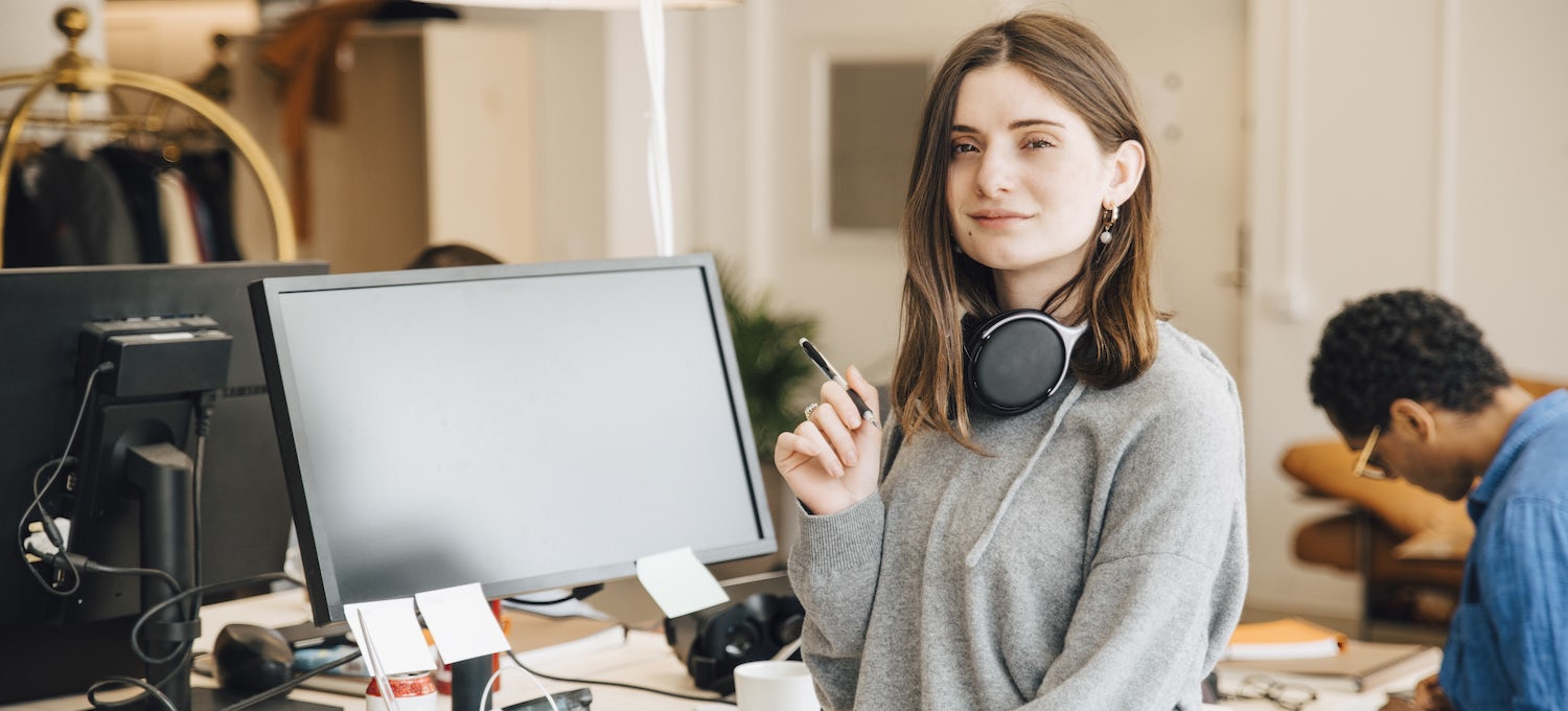 [Featured image] A young white woman in a gray long-sleeved top and over-the-ear headphones around her neck stands in front of a desktop computer. 