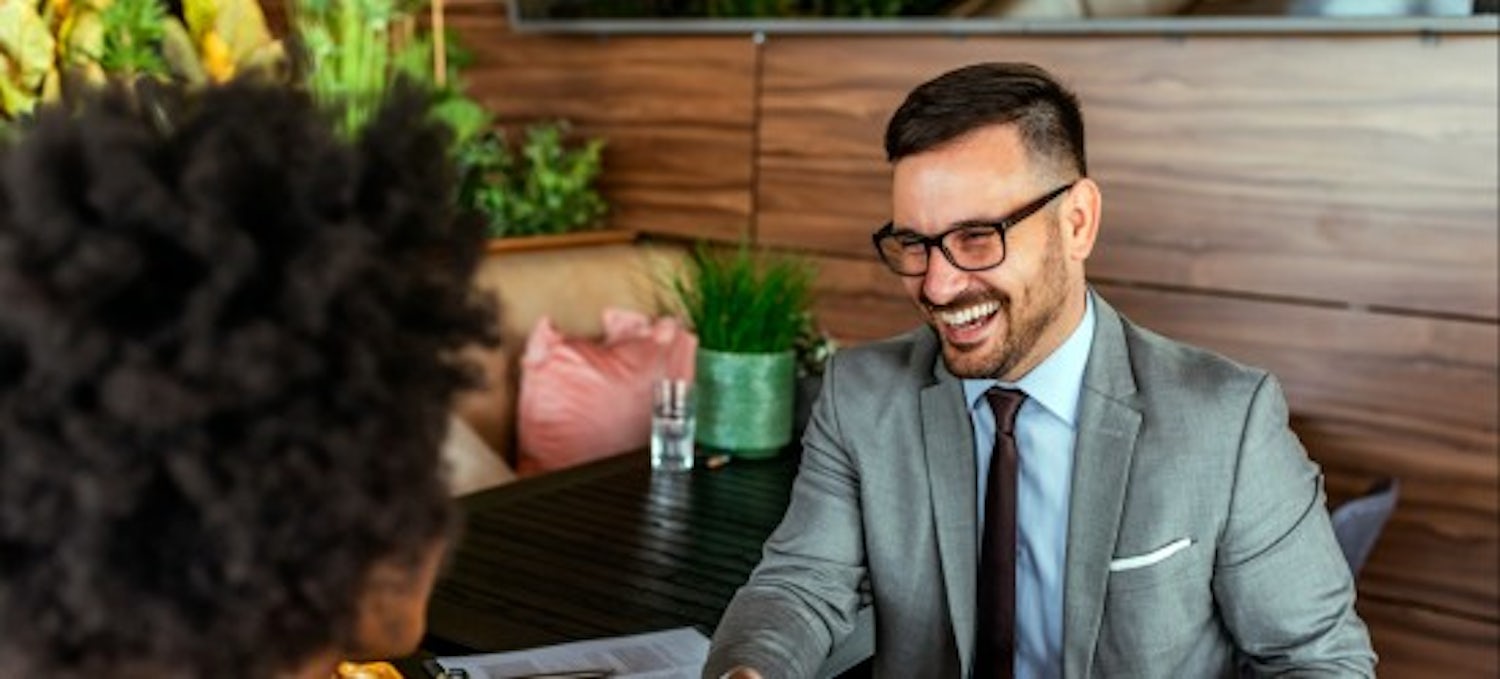 [Imagen destacada] Un hombre, con traje gris, camisa azul y corbata oscura, y gafas, está sentado en su mesa tras hablar con un posible empleado, en su puesto de recursos humanos. 