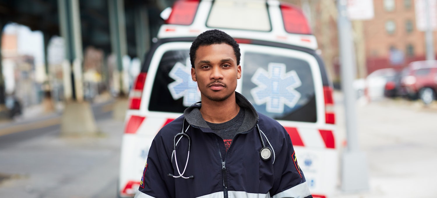 [Featured Image]: A man with short black hair and a mustache. He is wearing a black and white jacket. He has a stethoscope around his neck. He is standing in front of an ambulance. 